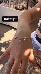 Sunlit hand wearing a delicate gold charm bracelet with a blue evil-eye bead and two sunburst/starburst charms, displayed over a tray of trinkets at an outdoor artisan market stall.