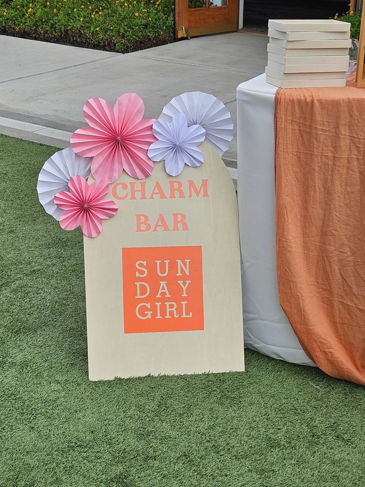 Arched cream event sign for a charm bar decorated with pink and white paper rosettes, standing on artificial grass beside a peach-draped table with stacked gift boxes