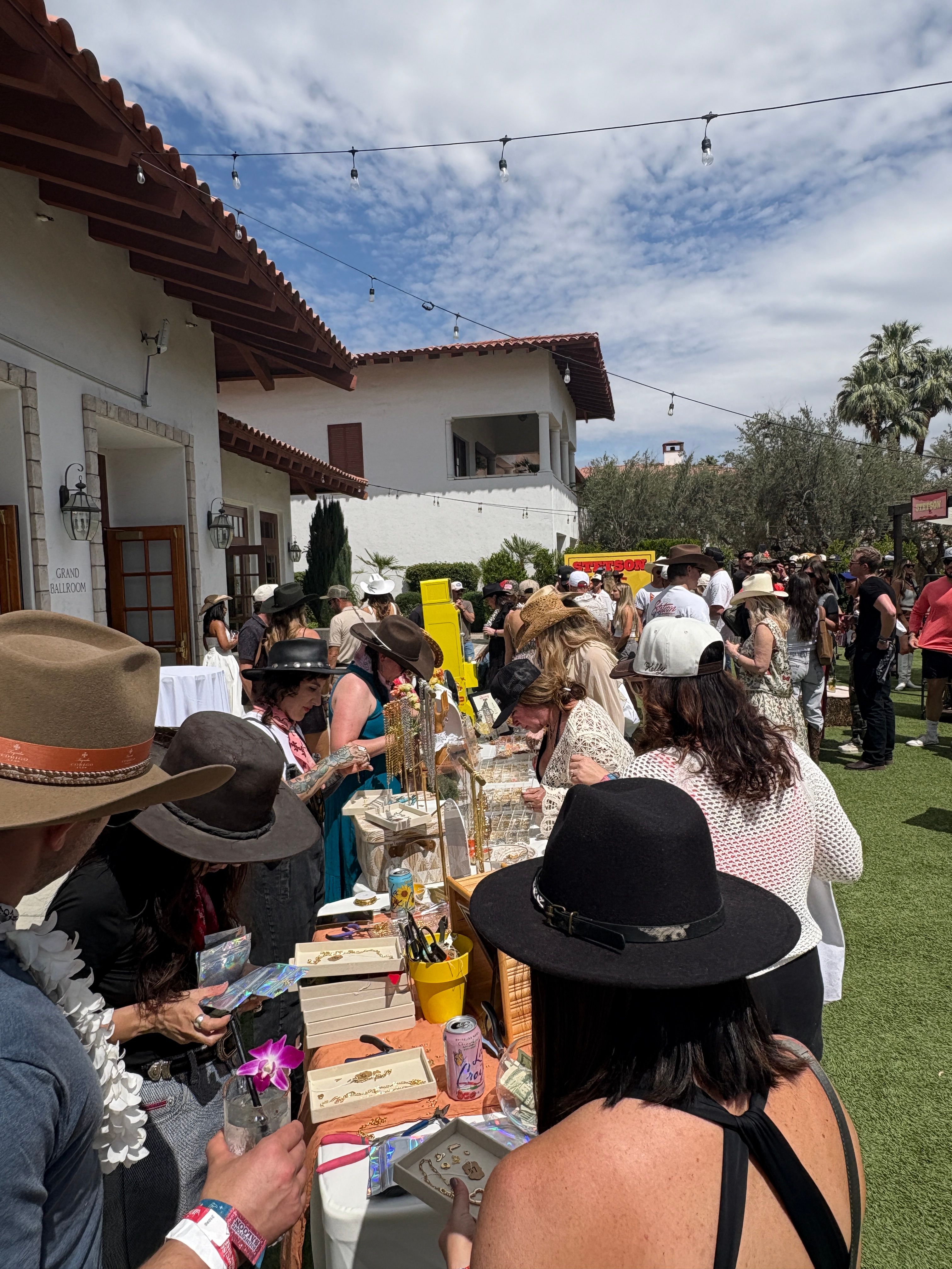 Sunny courtyard market at a Spanish-style villa — crowds in hats browsing jewelry and crafts under string lights with palm trees