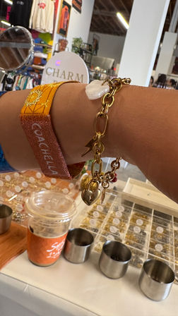 Close-up of a wrist wearing an orange Coachella fabric wristband and chunky gold charm bracelet with heart and pearl charms above a boutique jewelry counter