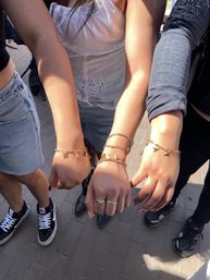 Three friends extending hands to show matching gold charm bracelets and rings on a sunlit city sidewalk, casual summer outfits with denim and sneakers.