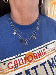 Close-up of neck and chest wearing layered silver charm necklaces (paw print, whale tail, clover, gemstone) over a blue California graphic T‑shirt with a hoop earring visible.