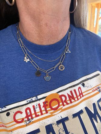 Close-up of neck and chest wearing layered silver charm necklaces (paw print, whale tail, clover, gemstone) over a blue California graphic T‑shirt with a hoop earring visible.