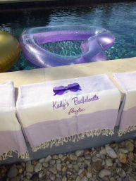 Backyard poolside scene with a purple glitter heart-shaped inflatable and gold float, a personalized lavender bachelorette towel draped over the pool edge with matching purple heart sunglasses on a pebble deck.