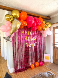 Bachelorette party backdrop in a home entryway: pink metallic fringe curtain with a “Veuve Before Vows” banner, oversized gold champagne foil balloon and vibrant garland of gold, orange, hot pink and blush balloons over a hardwood floor.