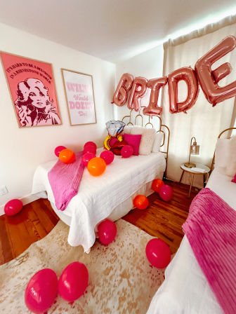 Cozy bedroom set up for a bachelorette party with rose-gold 'BRIDE' balloons and a diamond-ring foil balloon above white twin beds, pink throws and scattered pink and orange balloons.