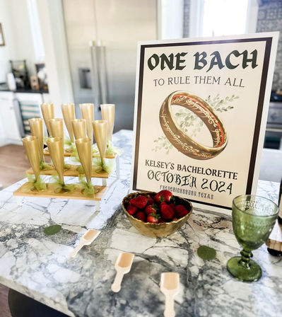 Bachelorette party display on a marble kitchen island: poster reading One Bach to Rule Them All with gold ring artwork, tiered gold champagne flutes tied with green bows, bowl of strawberries, green goblet and small wooden scoops.