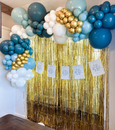 Festive indoor photo backdrop with teal, light blue, white and gold balloon garland framing a shimmering metallic gold fringe curtain — stylish party/photo booth decoration.