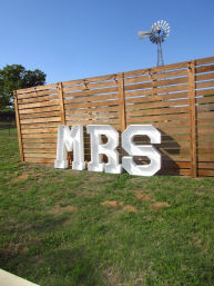 Outdoor rural scene with large white marquee letters spelling 'MRS' leaning against a rustic wooden slatted fence on a green lawn, a windmill and clear blue sky in the background