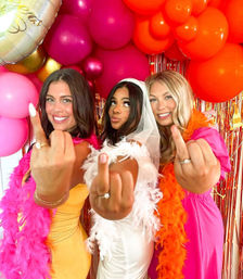 Three friends at a bachelorette party in front of pink and orange balloons, wearing feather boas and a bridal veil, playfully showing middle fingers to the camera.