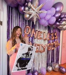 Smiling woman in a pink dotted dress at a purple-and-silver bachelorette backdrop, holding a poster with a black-and-white pop-star portrait and the words “ONE LESS LONELY GIRL,” surrounded by rose-gold letter balloons, a metallic star and clusters of purple and silver balloons.