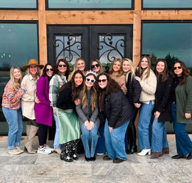 Smiling group of women in winter coats and boots posing outside a rustic wood-frame entrance with decorative black double doors — cheerful friends celebrating together.