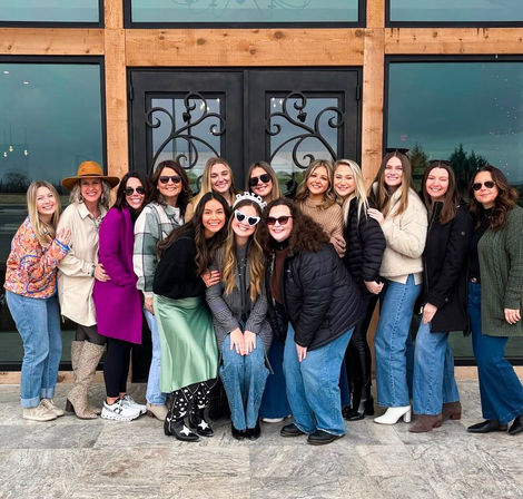 Smiling group of women in winter coats and boots posing outside a rustic wood-frame entrance with decorative black double doors — cheerful friends celebrating together.