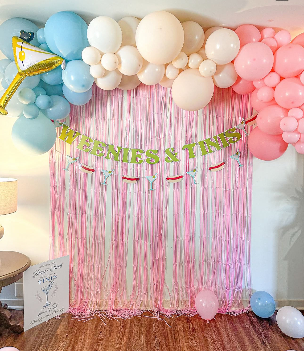 Playful party backdrop with pink fringe curtain, pastel blue, cream and pink balloon garland, “WEENIES & TINIS” banner and martini/hot dog garland above a wood floor with scattered balloons.