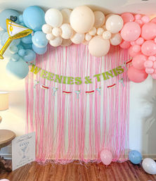 Playful party backdrop with pink fringe curtain, pastel blue, cream and pink balloon garland, “WEENIES & TINIS” banner and martini/hot dog garland above a wood floor with scattered balloons.