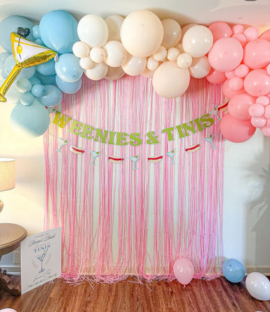 Playful party backdrop with pink fringe curtain, pastel blue, cream and pink balloon garland, “WEENIES & TINIS” banner and martini/hot dog garland above a wood floor with scattered balloons.