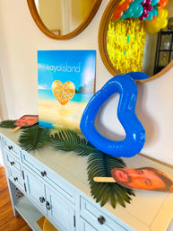 Beach-themed party vignette on a pale blue console with tropical palm leaves, a blue inflatable heart float, glittery heart beach poster, face-cutout photo props and round mirror reflecting colorful balloons — Fredericksburg, Texas