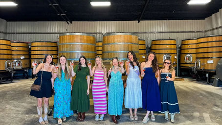 Nine women in colorful summer dresses smiling and toasting with wine glasses in a winery barrel room lined with large oak fermentation tanks.