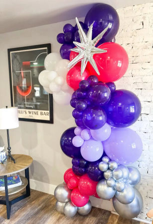 Indoor party balloon garland with purple, lavender, hot pink, white and metallic silver clusters, topped by a silver star foil balloon against a white brick wall beside a wooden side table and lamp.