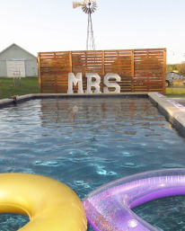 Backyard outdoor pool with gold and glittery purple inflatables in the foreground and large marquee letters spelling MRS on a wooden slat fence, rustic windmill and barn visible in the background.