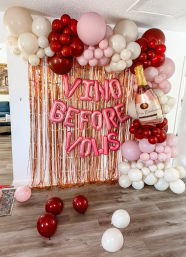 Indoor rose-gold fringe photo backdrop with pink metallic "VINO BEFORE VOWS" letter balloons, oversized champagne bottle balloon and pink, red and white balloon garlands for a bachelorette party