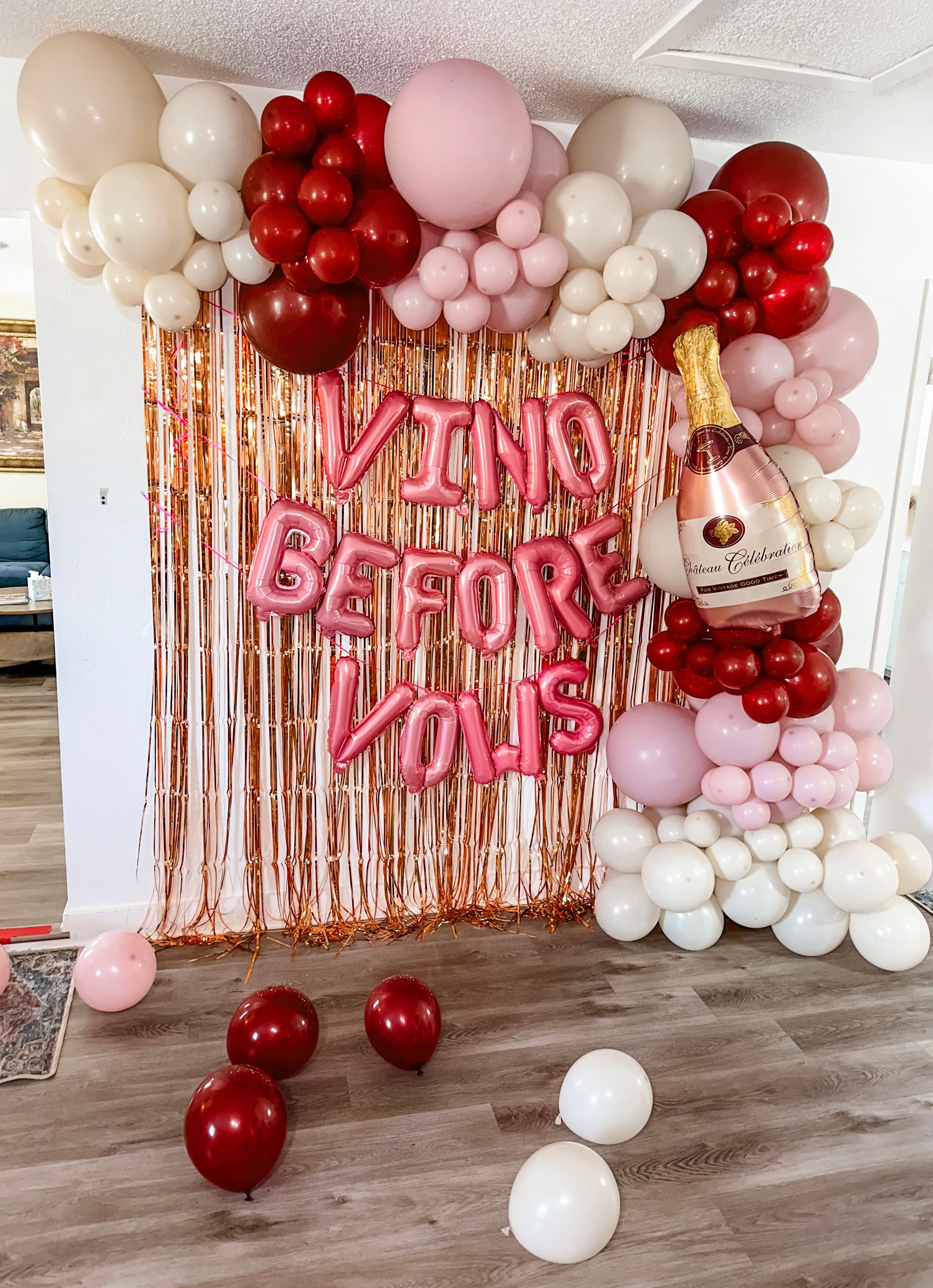 Indoor bachelorette party backdrop in a living room: rose-gold fringe curtain with pink foil letters reading 'VINO BEFORE VOWS', pink, red and ivory balloon arch and oversized champagne bottle balloon.
