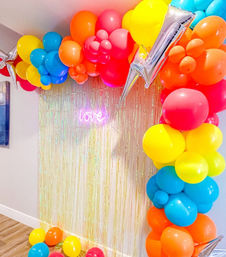Colorful rainbow balloon arch framing an iridescent fringe photo backdrop with a pink neon "love" sign, silver star foil balloons and scattered balloons on a hardwood floor — indoor party decor