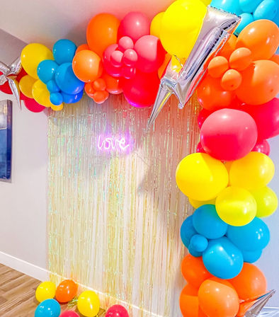 Colorful rainbow balloon arch framing an iridescent fringe photo backdrop with a pink neon "love" sign, silver star foil balloons and scattered balloons on a hardwood floor — indoor party decor