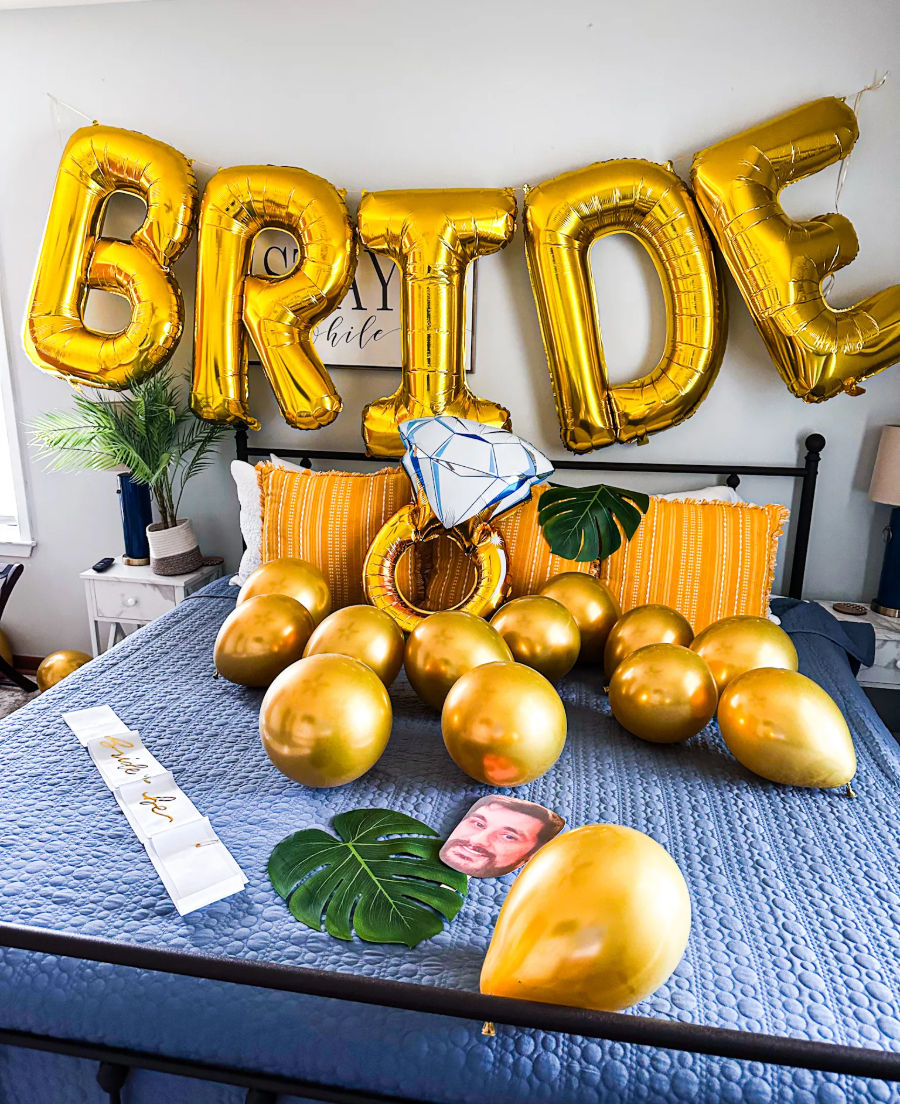 Bedroom staged for a bachelorette: large gold BRIDE foil balloons strung above a bed, scattered gold latex balloons, oversized inflatable diamond ring prop, tropical leaves and yellow pillows on a blue quilted comforter.