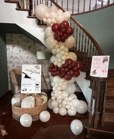 Curved wooden staircase foyer decorated with a cream and burgundy balloon garland, wine-themed welcome signs, cowhide ottoman and scattered balloons