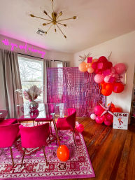 Vibrant pink dining room party setup with glass table, hot-pink velvet chairs, patterned pink rug, sequin photo backdrop, pink-orange balloon garland, modern brass chandelier and neon wall sign.