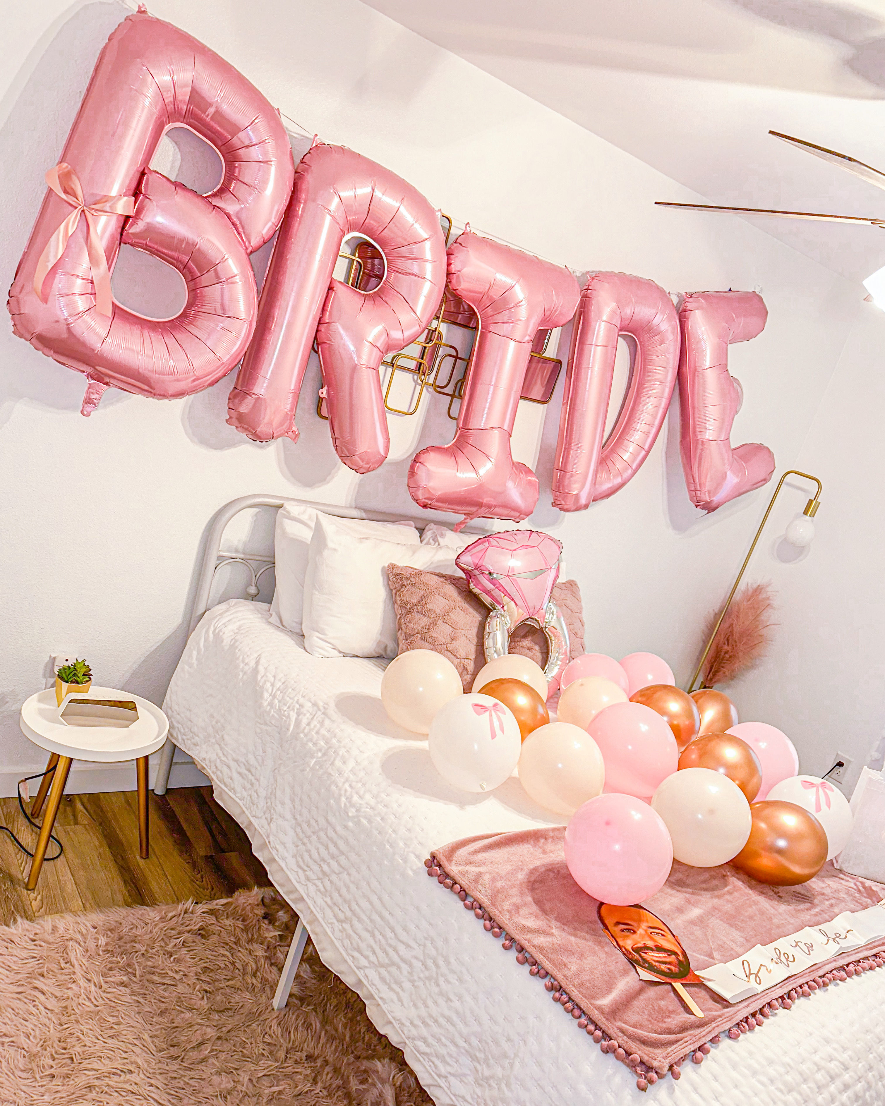 Rose-pink 'BRIDE' foil balloons above a white bed styled with pink and rose-gold balloons, ring-shaped balloon, textured pillows, plush pink throw and bedside table with succulent — bachelorette/bridal party decor.