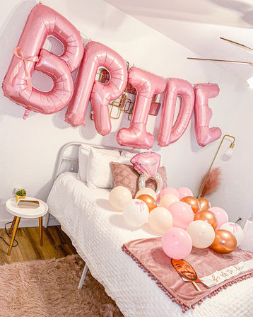 Rose-pink 'BRIDE' foil balloons above a white bed styled with pink and rose-gold balloons, ring-shaped balloon, textured pillows, plush pink throw and bedside table with succulent — bachelorette/bridal party decor.