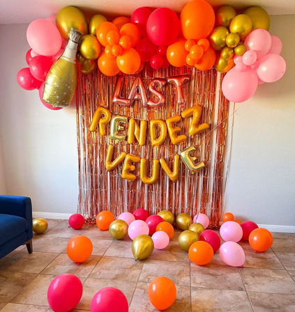 Indoor party backdrop with rose-gold fringe curtain, gold letter balloons spelling a celebratory message, bright pink, orange and metallic gold balloon garland, a foil champagne bottle balloon, and scattered balloons on a tiled living-room floor.