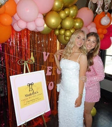 Two smiling women pose at an indoor bridal shower celebration beneath a pink, gold and orange balloon garland and metallic fringe backdrop, one in a white textured dress and the other in a pink sequin mini dress beside a decorative easel sign.