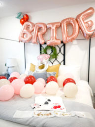 Festive bridal shower bedroom setup with rose-gold "BRIDE" balloons above a bed scattered with pink, red and white balloons, ring-shaped foil balloon, decorative pillows, folded shirt, sunglasses and a "bride to be" sash.