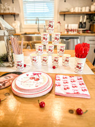 Cherry-themed party display on a home kitchen island — pyramid of paper cups with cherry print, matching plates and napkins, red plastic utensils and striped straws on a wood countertop.