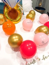 Bachelorette bedroom setup with pink, orange, red and metallic gold logo-printed balloons, a large gold ring-shaped foil balloon and a bride-to-be sash on a white bed.