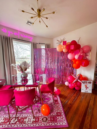 Vibrant pink-themed dining room party setup with glass table and hot-pink velvet chairs on a patterned pink rug, disco-ball vase centerpiece, sequin photo backdrop and pink-orange balloon garland under a brass chandelier.