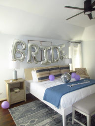 Master bedroom decorated for a bridal celebration: large silver foil "BRIDE" balloons over a white bed with a blue throw, purple balloons, disco ball, nightstands and ceiling fan.