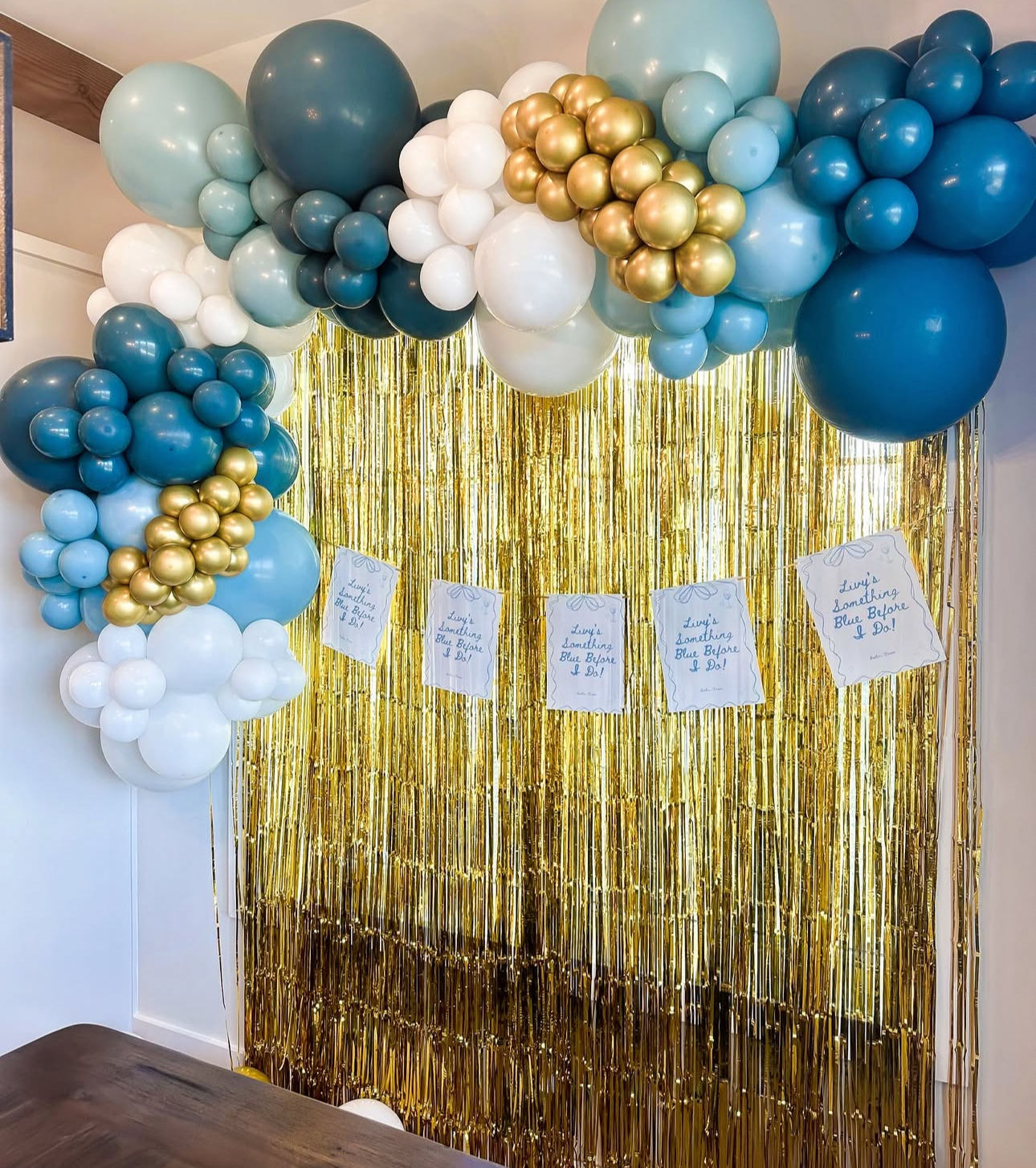 Festive indoor photo backdrop with teal, blue, white and metallic gold balloon garland arching over a shiny gold tinsel fringe curtain and small paper signs