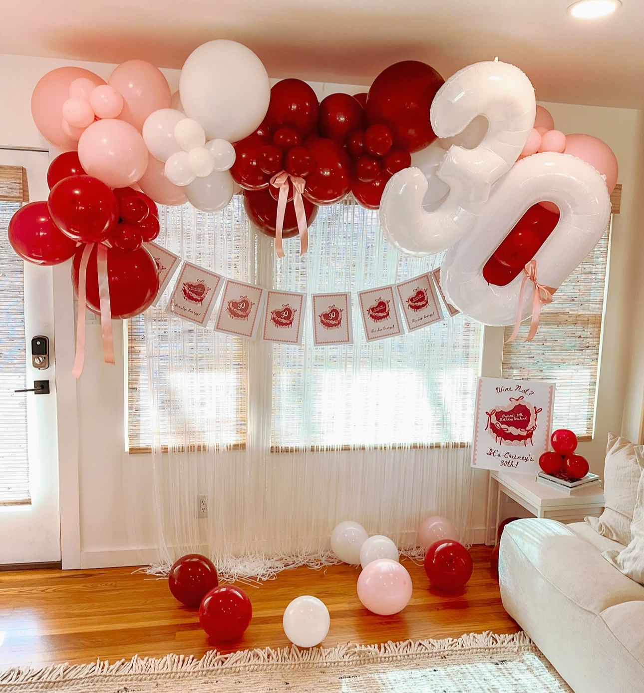 Festive pink, red and white balloon arch with oversized white '30' foil number and birthday banner in a cozy living room, balloons scattered on hardwood floor