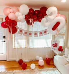 Festive pink, red and white balloon arch with oversized white '30' foil number and birthday banner in a cozy living room, balloons scattered on hardwood floor