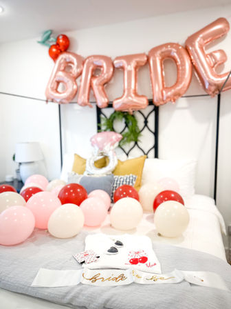 Bright home bachelorette bedroom scene with rose-gold "BRIDE" balloons over a bed strewn with pink, red and white balloons, gold pillows, and a "bride-to-be" sash and cherry-themed robe.