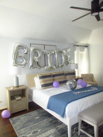 Bright modern bedroom styled for a bachelorette: silver 'BRIDE' foil balloons above bed, disco ball, purple balloons and blue throw