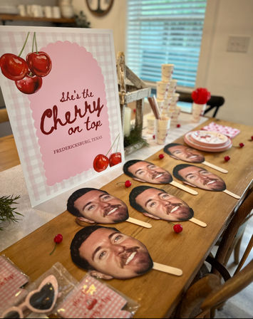 Cherry-themed party table in Fredericksburg, Texas with a pink “She’s the Cherry on Top” sign, stacked paper cups and plates, scattered faux cherries and multiple smiling face paddle photo props on sticks arranged on a wooden table.