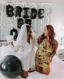 Two friends laughing on a bed at a bachelorette party in a bedroom — one wearing a white veil and shirt holding a 'bride' goblet, the other in a gold sequin mini dress with balloons and silver fringe backdrop