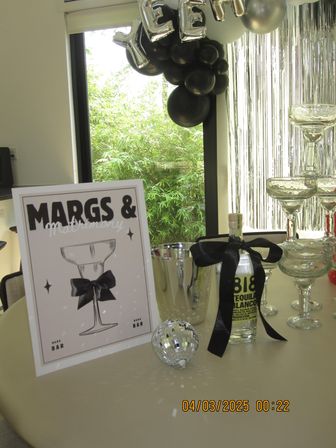 Sunlit indoor party bar by a window with bamboo outside — framed cocktail sign, stacked coupe glasses, tequila bottle tied with a black ribbon, silver ice bucket, mini disco ball, black balloons and silver fringe backdrop.
