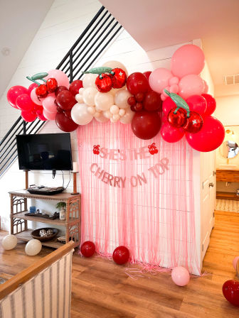 Cherry-themed home party backdrop with pink, red and cream balloon arch, cherry foil balloons, pink fringe curtain and "She's the cherry on top" banner