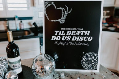 Bachelorette party poster on a kitchen counter in Fredericksburg, Texas that reads Til Death Do Us Disco with a skeletal hand heart graphic, mirrored disco balls, scattered confetti, and a bottle of red wine.